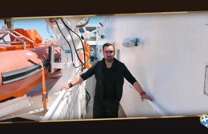 Man standing on a white ship deck beside an orange lifeboat, wearing black clothing and sunglasses, with railing on either side.
