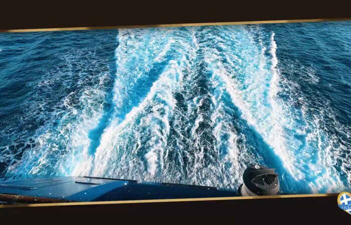 View from a boat looking back at a bright blue sea, white wake trails stretching into the distance, a logo in the bottom-right corner.