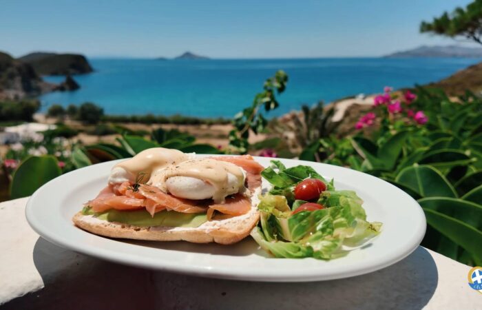 Plate with smoked-salmon Eggs Benedict and side salad on a white table, ocean view in the background.