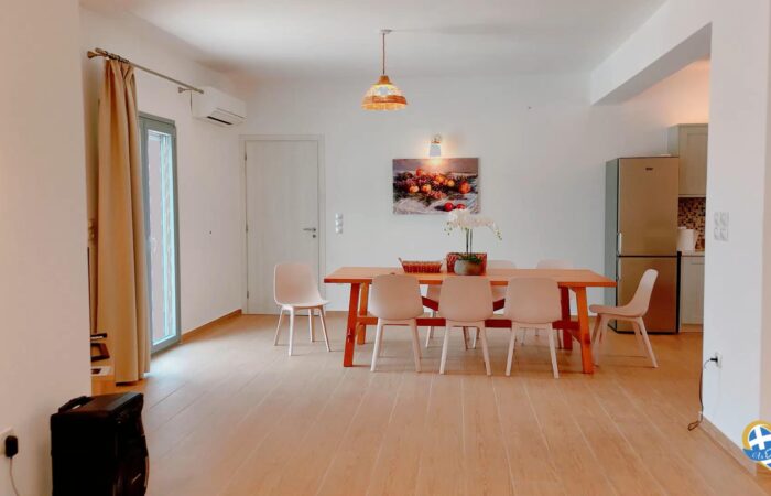 Open-plan dining area with a wooden table, six white chairs, a pendant light, wall art, and a fridge visible in the kitchen.