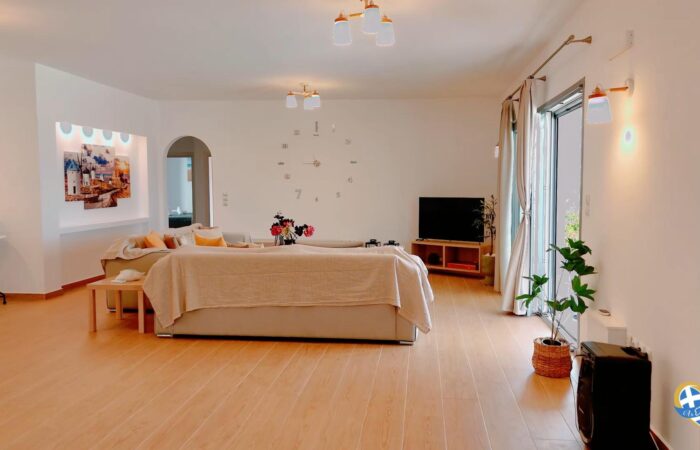 Bright living room with a beige sectional sofa, colorful cushions, and a TV on a wooden stand facing large glass doors.
