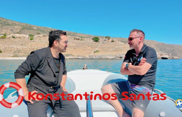 Two men sit on a small boat near a rocky coastline, wearing sunglasses, with clear blue water and sky; red text overlay reads 'Konstantinos Santas'. A life ring is on the left and a logo appears bottom-right.