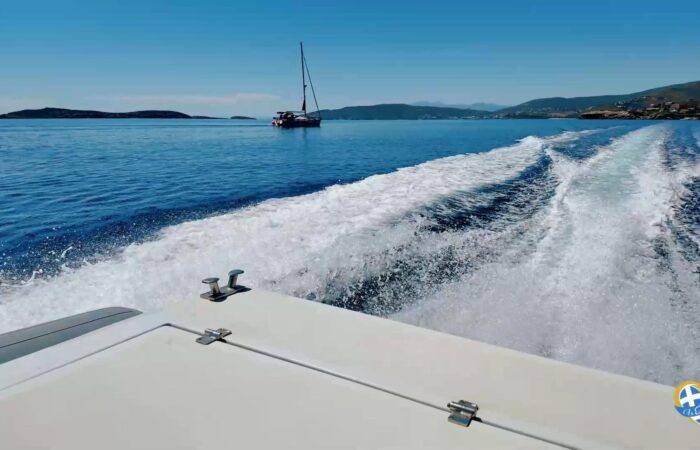 View from a speeding boat with a wake in the foreground, a sailboat ahead on calm blue water, and a coastline in the distance.
