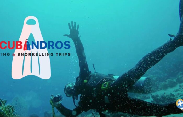 Underwater scene promoting Scubandros: a diver swimming with a camera, logo text 'SCUBANDROS DIVING & SNORKELLING TRIPS' overlaid on the left, and a heart-shaped Greek flag badge bottom right.