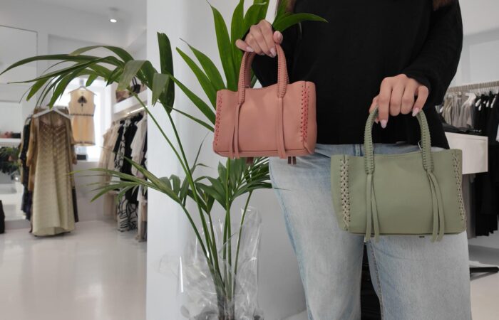 A person in a black top and jeans holds two braided-handled handbags, one pink and one light green, in a bright clothing boutique.