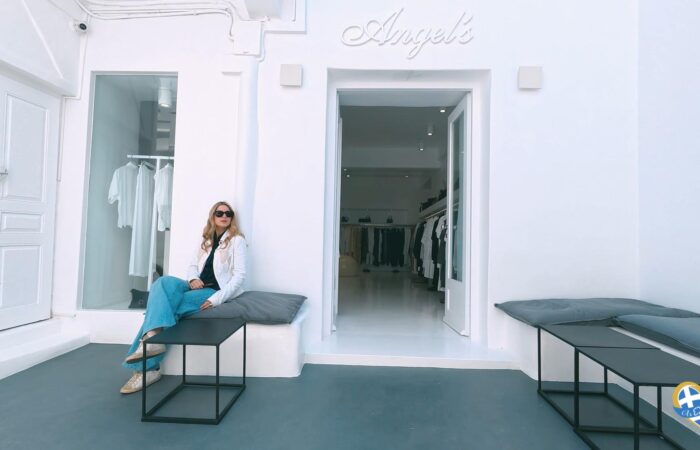 Woman in a white jacket and blue jeans sits on a bench outside a white clothing boutique with clothes visible through the open door.