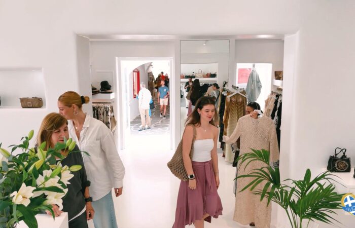 Shoppers browse clothes in a bright white boutique with handbag displays and plants in the foreground.