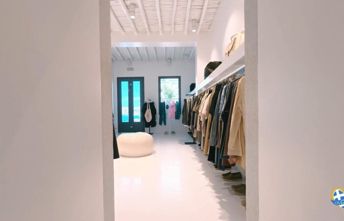Clothing store interior with racks along the right side, white floor and ceiling, and a blue door at the back; a round white seating area in the center.