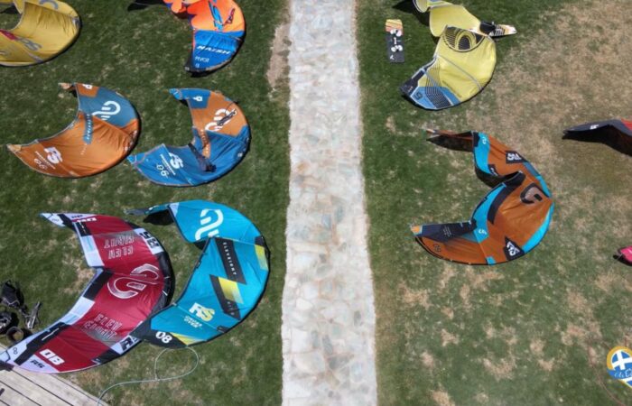 Colorful kites laid out on a grassy field around a central stone path, seen from above, likely preparing for a kiteboarding event; bottom right shows a heart-shaped blue-and-yellow logo.