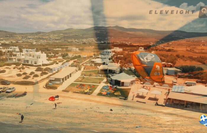 Aerial view of a coastal resort with white modern villas, a sandy beach, and water-sport gear including an orange parasail wing overhead.