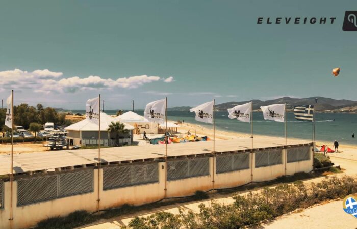 Beachside promenade along a sandy beach with white flag banners, tents, and people by the shore under a clear blue sky.