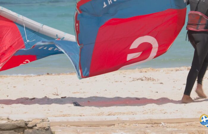 Person in a black wetsuit stands on a sandy beach with a red-blue windsurfing sail near the water. A heart-shaped logo is in the bottom right corner.