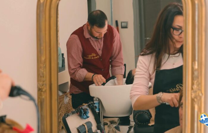 Barber in a burgundy vest washes a client’s hair at a salon sink, with a stylist in the foreground nearby.