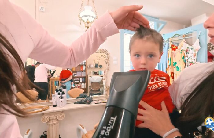 Child in a red shirt sits on a chair while a stylist trims his hair in a bright boutique salon, with two adults nearby watching or assisting.