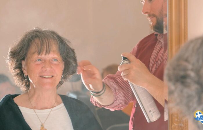 Smiling older woman seated in a salon chair as a hairdresser sprays her hair with a can, seen in the mirror behind them.