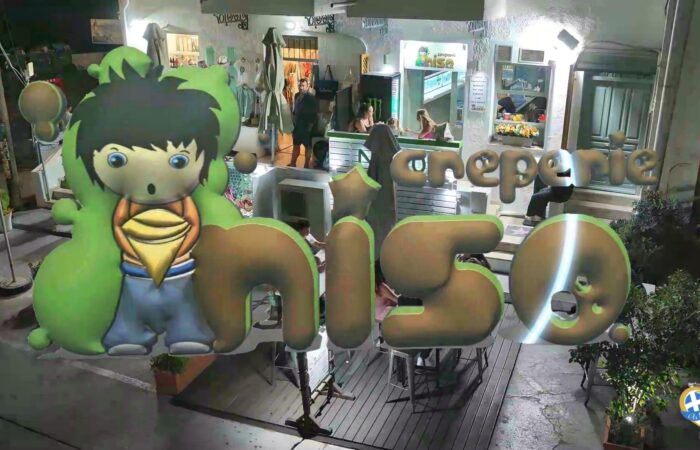 Nighttime storefront with a giant green cartoon boy statue and oversized metallic letters spelling the shop name, with customers visible inside.