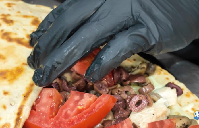 Gloved hand placing chopped tomatoes and olives into a flatbread wrap with meat filling.