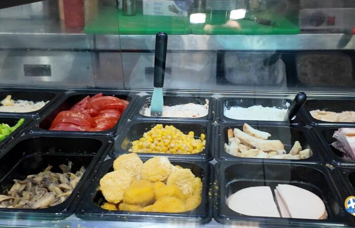 Self-serve deli counter with multiple trays of food, including corn, sliced tomatoes, fried chicken, and mushrooms, with a serving knife in the center.