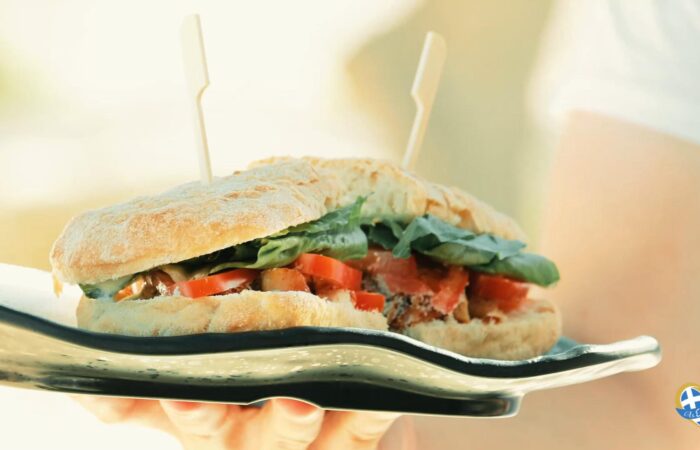 Close-up of a rustic sandwich filled with greens and tomato, held on a glossy plate with two wooden skewers piercing the bread.