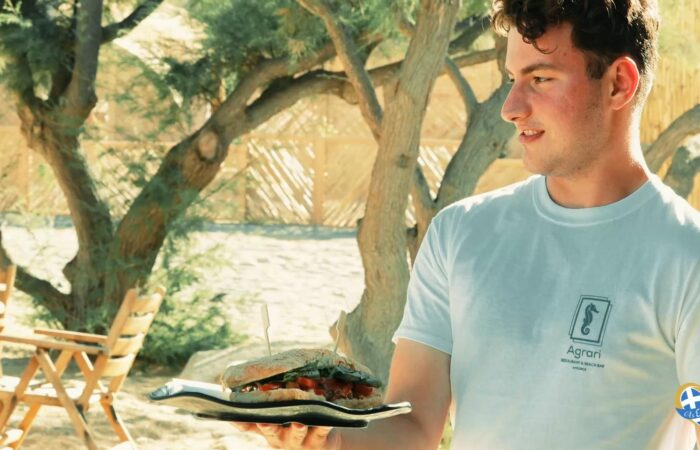 Man wearing a light blue shirt holds a plate with a tall, stacked burger outdoors, with wooden chairs and trees in the background.
