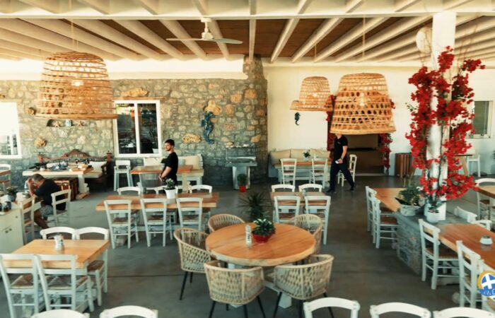 Bright cafe interior with a stone wall, wooden tables, and white chairs, large woven pendant lights, and red flowering vines.