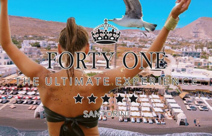 Back view of a woman with arms raised overlooking a Santorini beach club and white buildings, with promotional text 'FORTY ONE' and 'THE ULTIMATE EXPERIENCE'.