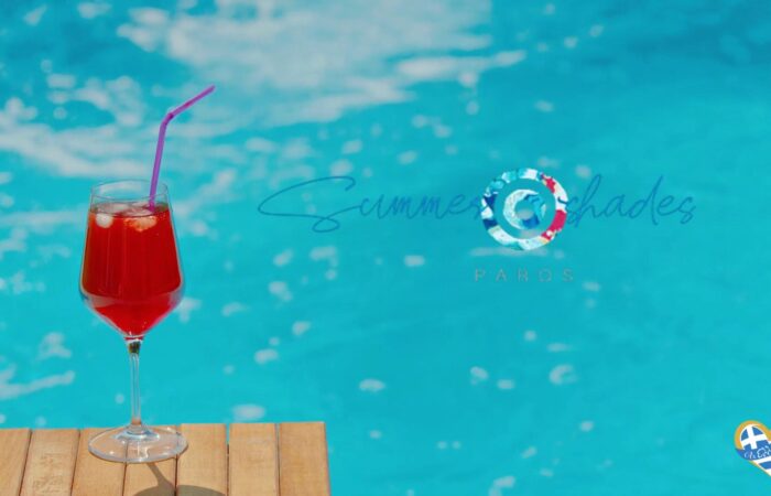 Red cocktail in a stemmed glass with a purple straw, placed on a wooden surface beside a bright blue pool behind. The 'Summer Shades' logo is visible in the background.