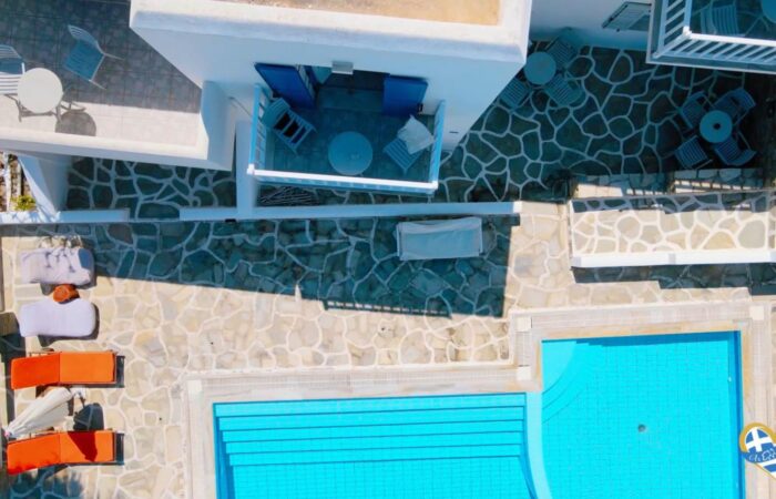 Aerial view of a white-walled resort courtyard with a bright blue rectangular pool, stone-paved decks, orange lounge chairs, and white tables/chairs on upper terraces.