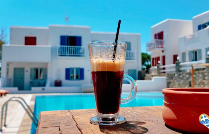Glass of iced coffee with a black straw on a wooden poolside table, blue pool and white modern buildings in the background.