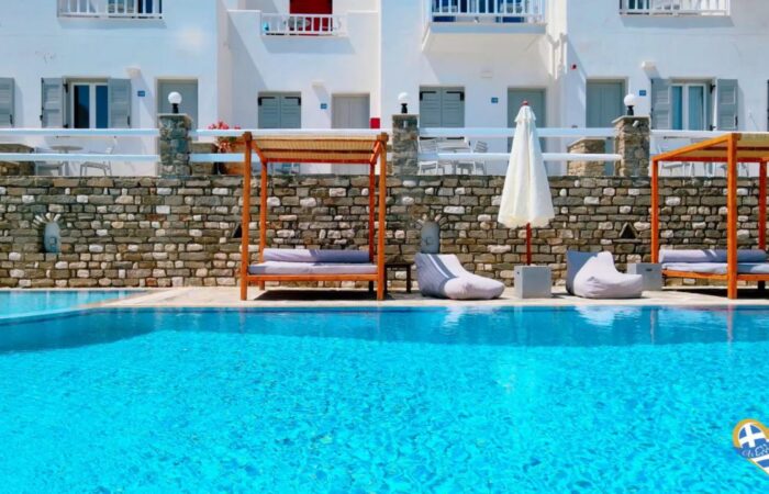 Crystal blue swimming pool with wooden cabanas and a stone wall backdrop, white buildings, and lounge seating along the pool deck.