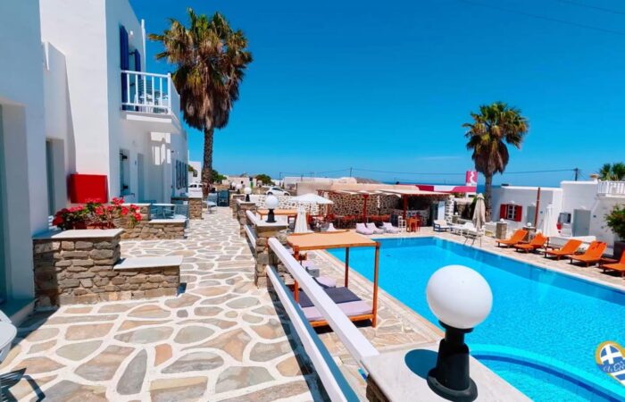 White-washed hotel courtyard with a bright blue pool, stone walkways, and orange loungers under clear skies.
